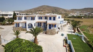 A two-story building with a white facade and blue accents, surrounded by palm trees and a well-maintained lawn. - Akti Aegeou Hotel (Tinos)