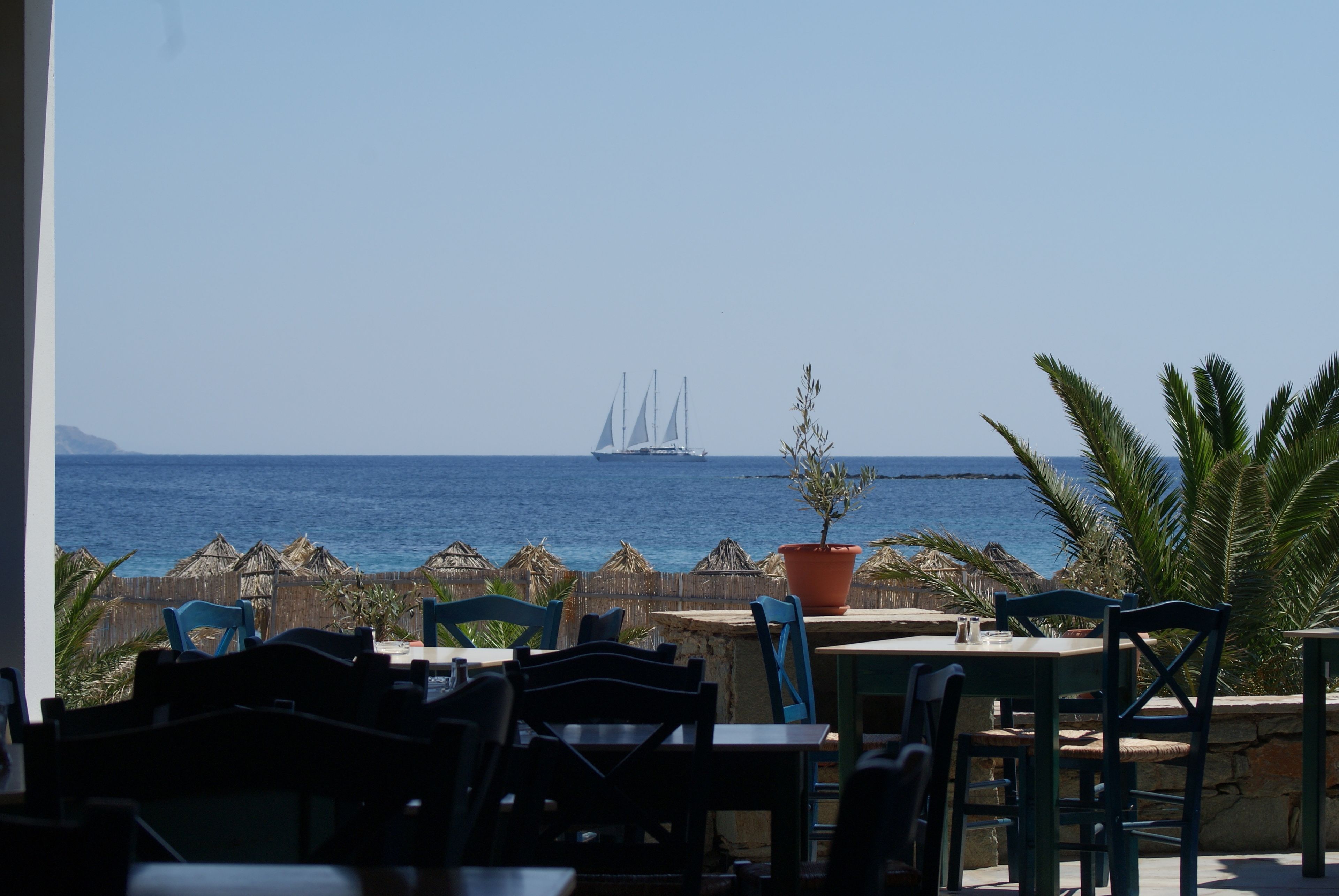An outdoor seating area with tables and chairs, overlooking the sea and a distant sailboat.