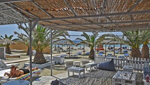 A covered outdoor area with lounge chairs and umbrellas, overlooking a beach with palm trees and a clear sky. - Akti Aegeou Hotel (Tinos)