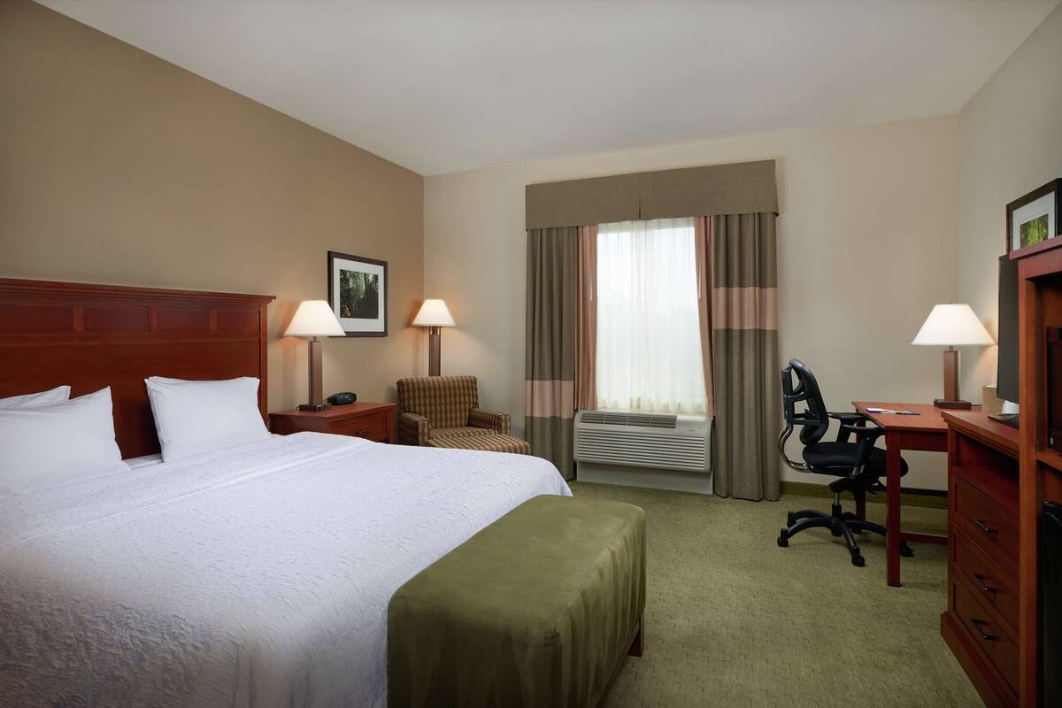 A hotel room with a large bed, two bedside tables with lamps, a chair, a desk with a chair, and an air conditioning unit.