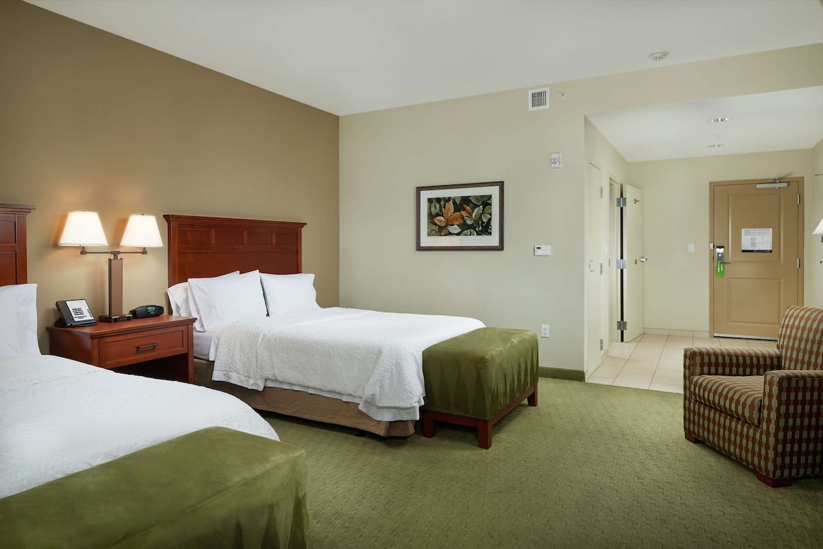 A hotel room with two beds, a wooden headboard, a green ottoman, a chair, and a painting on the wall.