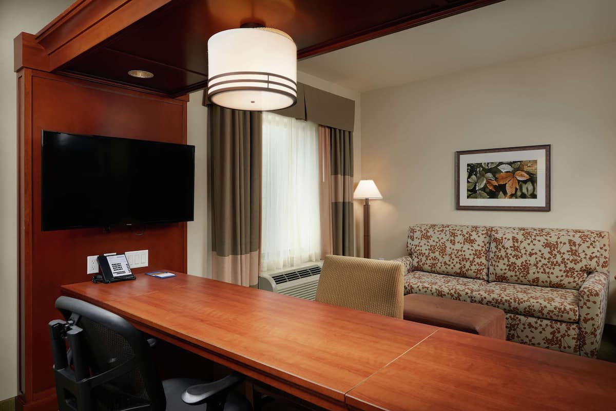 A hotel room with a wooden desk, a flat-screen TV, a sofa, a lamp, and a framed picture on the wall.