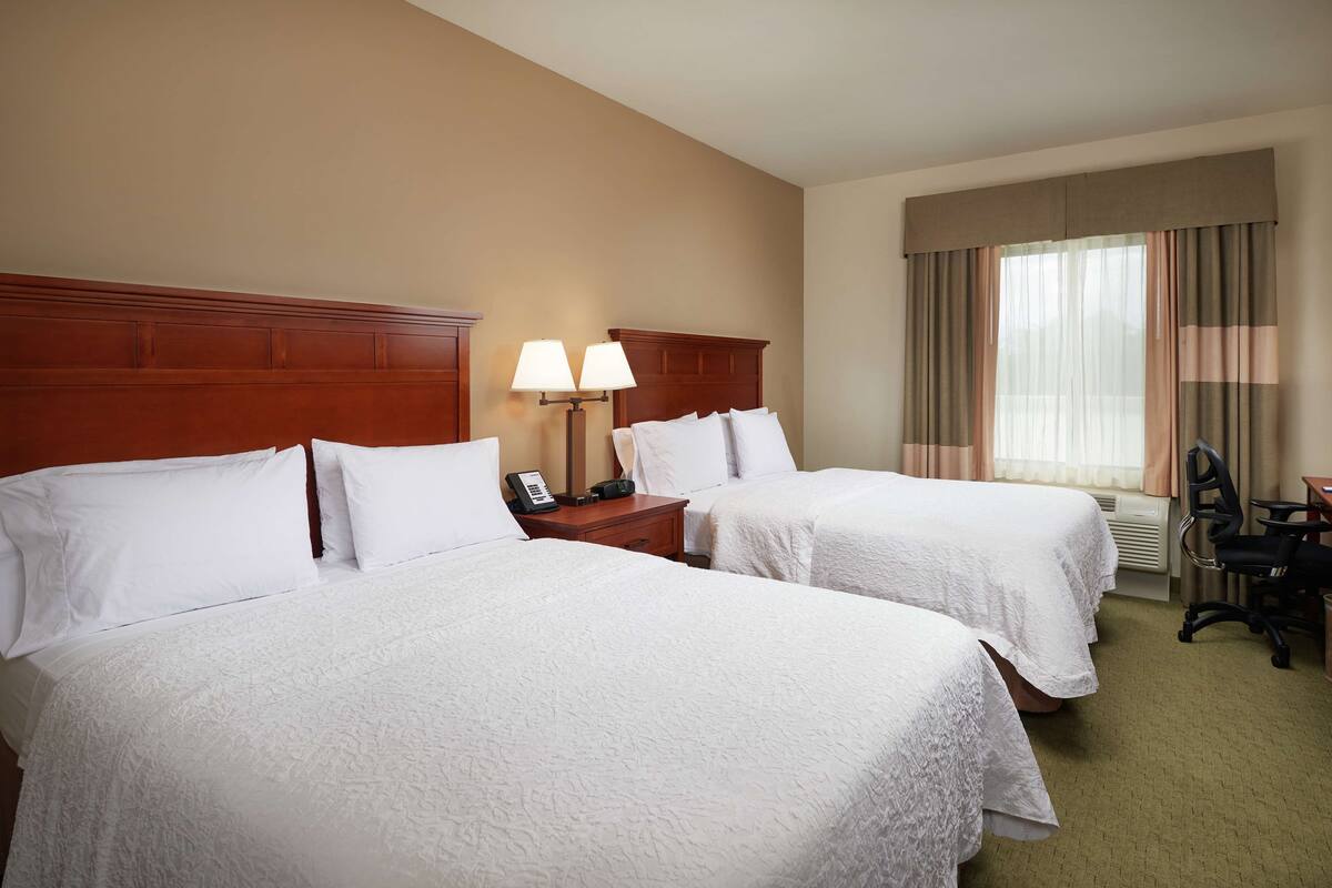 A hotel room with two beds, a nightstand with a lamp, a desk with a chair, and a window with curtains.