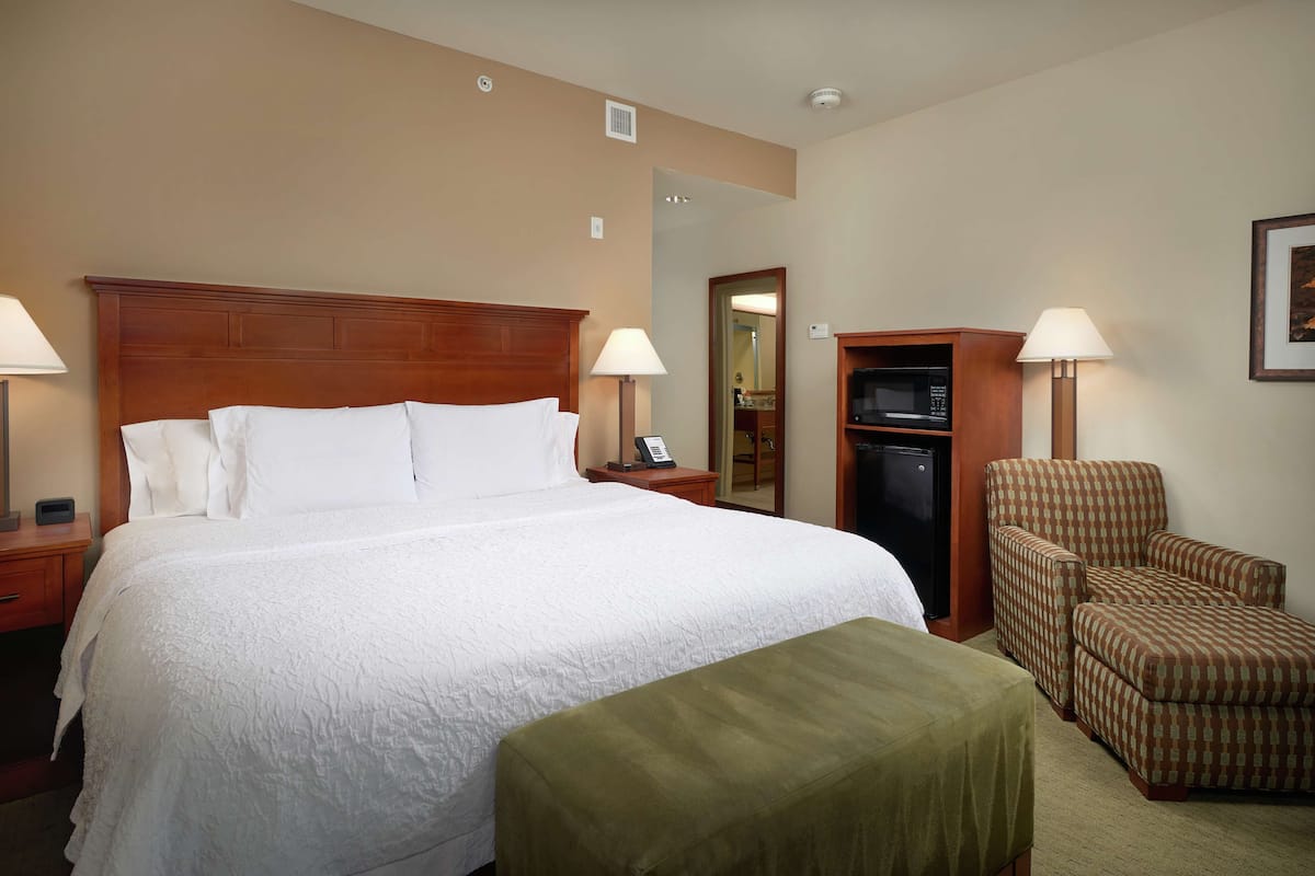 A hotel room with a large bed, two bedside lamps, a green ottoman, a checkered armchair, a wardrobe, and a mirror.