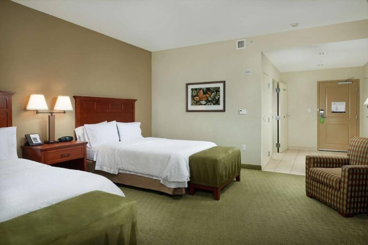 Habitación de hotel con dos camas, una silla, una mesita de noche con lámpara, un cuadro enmarcado en la pared y una puerta que da a otra habitación.