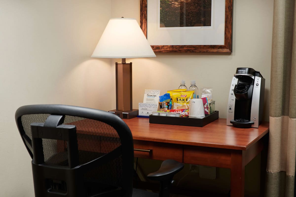 A hotel room with a wooden desk, a black mesh chair, a coffee maker, a lamp, and a framed picture on the wall.