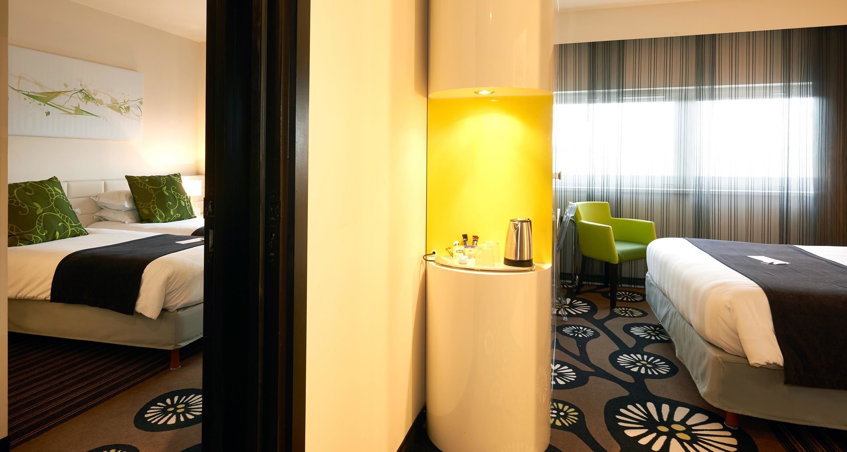 Habitación de hotel con dos camas, una mesita de noche con artículos de aseo, una silla verde y un ventanal con cortinas.