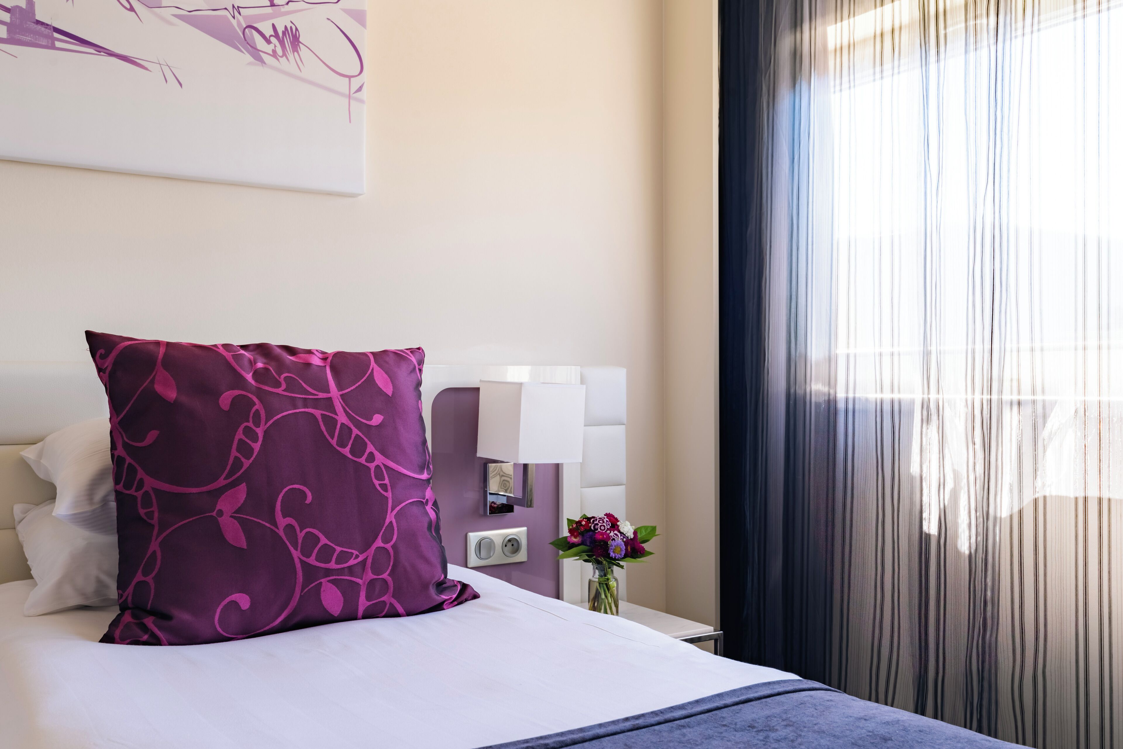 Een netjes opgemaakt bed met een paars kussen, een nachtkastje met een lamp en een vaas met bloemen.