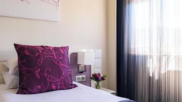 Een netjes opgemaakt bed met een paars kussen, een nachtkastje met een lamp en een vaas met bloemen.