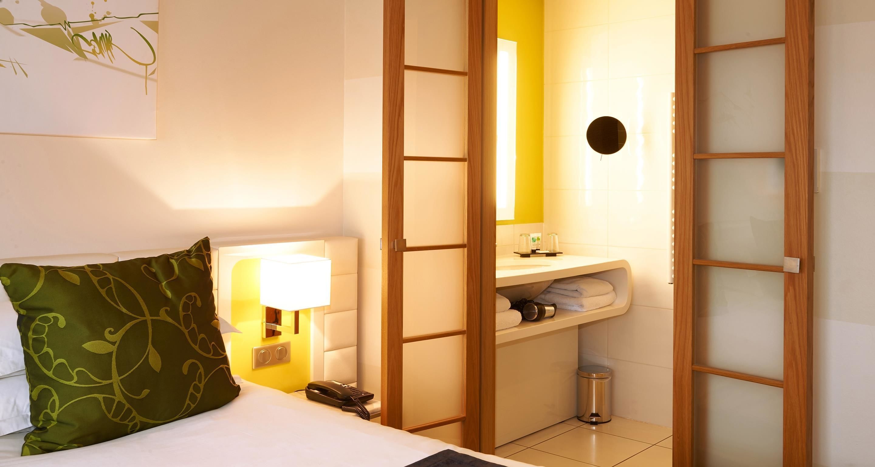 Habitación de hotel con cama, lámpara de noche, espejo, lavabo y cubo de basura.