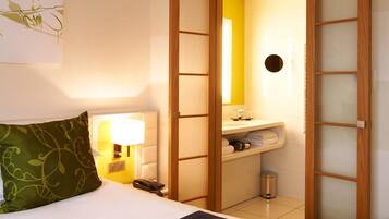 Habitación de hotel con cama, lámpara de noche, espejo, lavabo y cubo de basura.