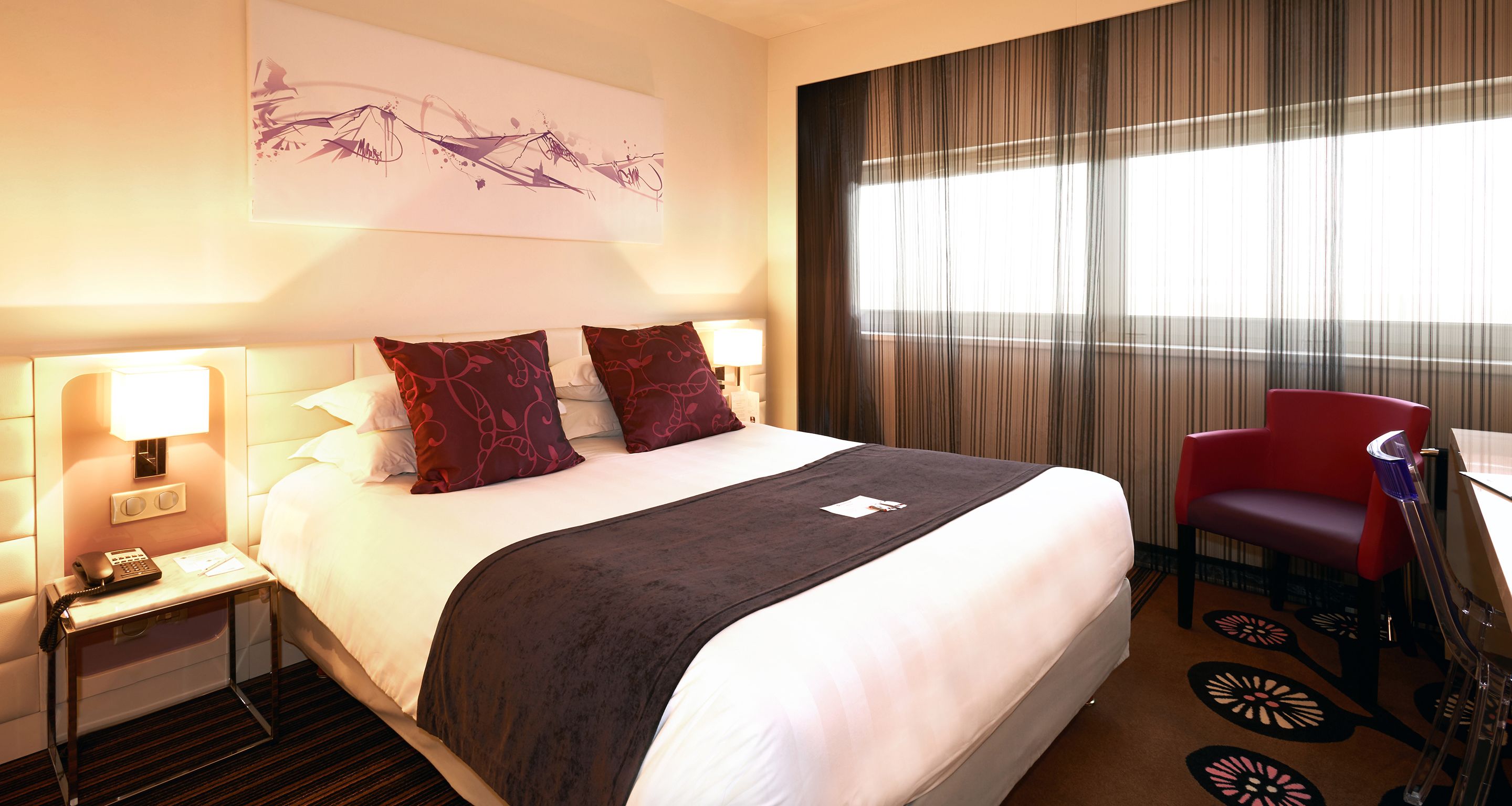 Habitación de hotel con una cama grande, dos lámparas de noche, un escritorio con teléfono, una silla roja y un ventanal con cortinas.