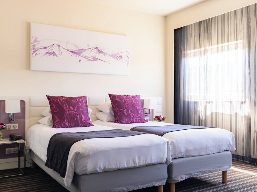 A hotel room with two beds, a bedside table with a lamp, a phone, and a vase with flowers.