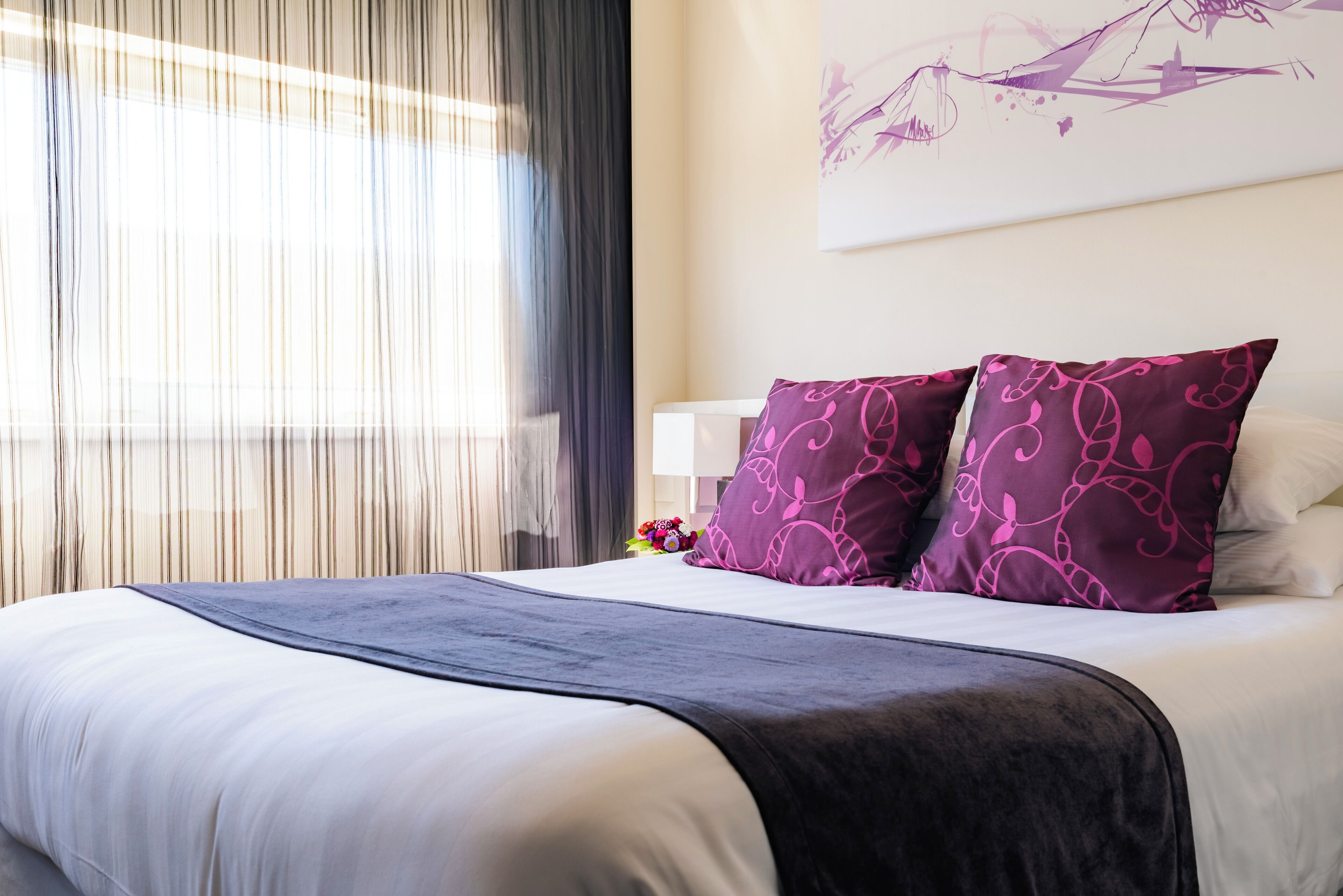 A neatly made bed with a white and dark gray bedspread, two purple pillows with a leaf pattern, and a wall-mounted artwork above.
