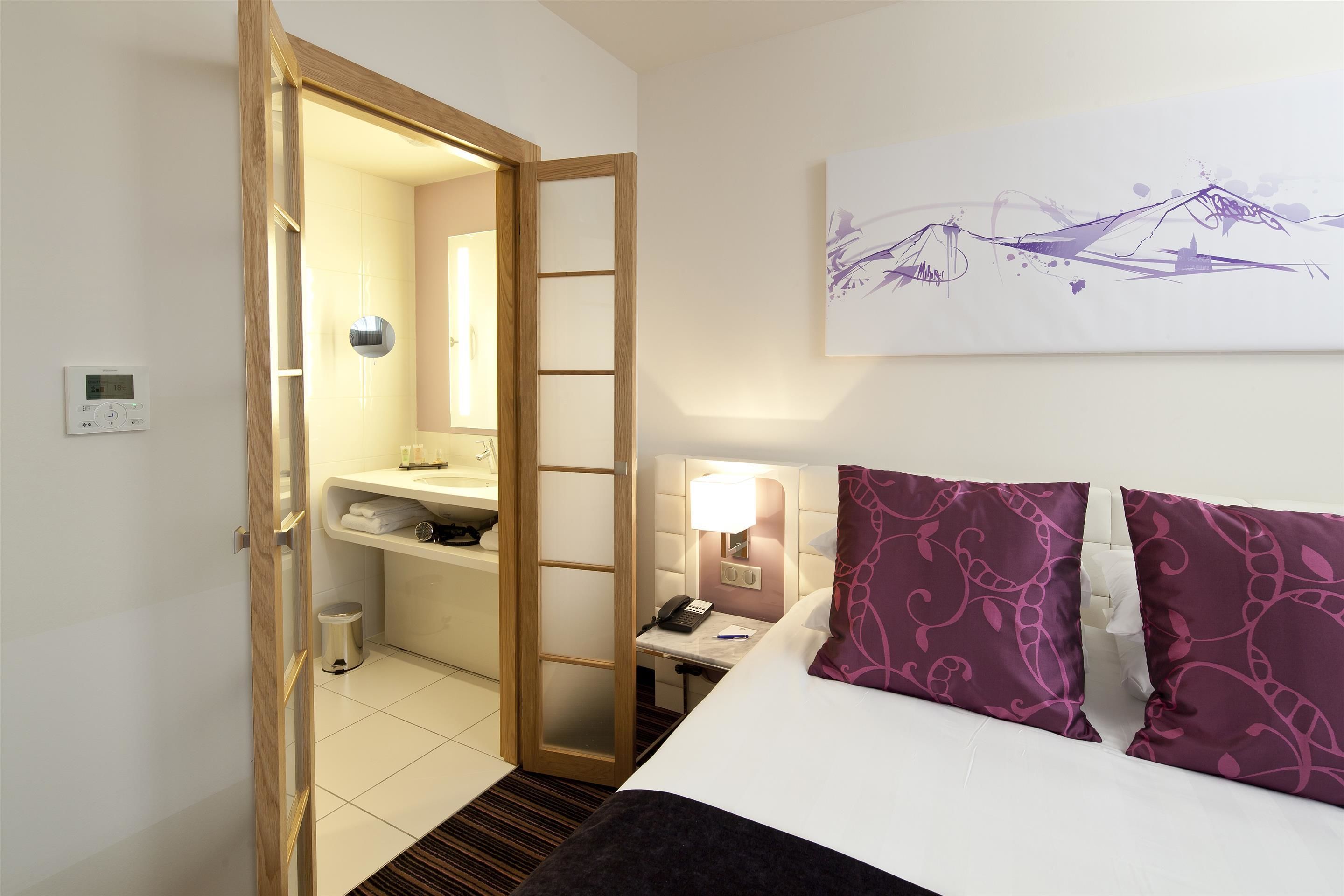 A hotel room with a bed, a bedside table with a lamp, a phone, and a small trash bin.