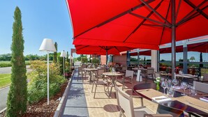 An outdoor seating area with red umbrellas, tables, and chairs, overlooking a road and greenery.