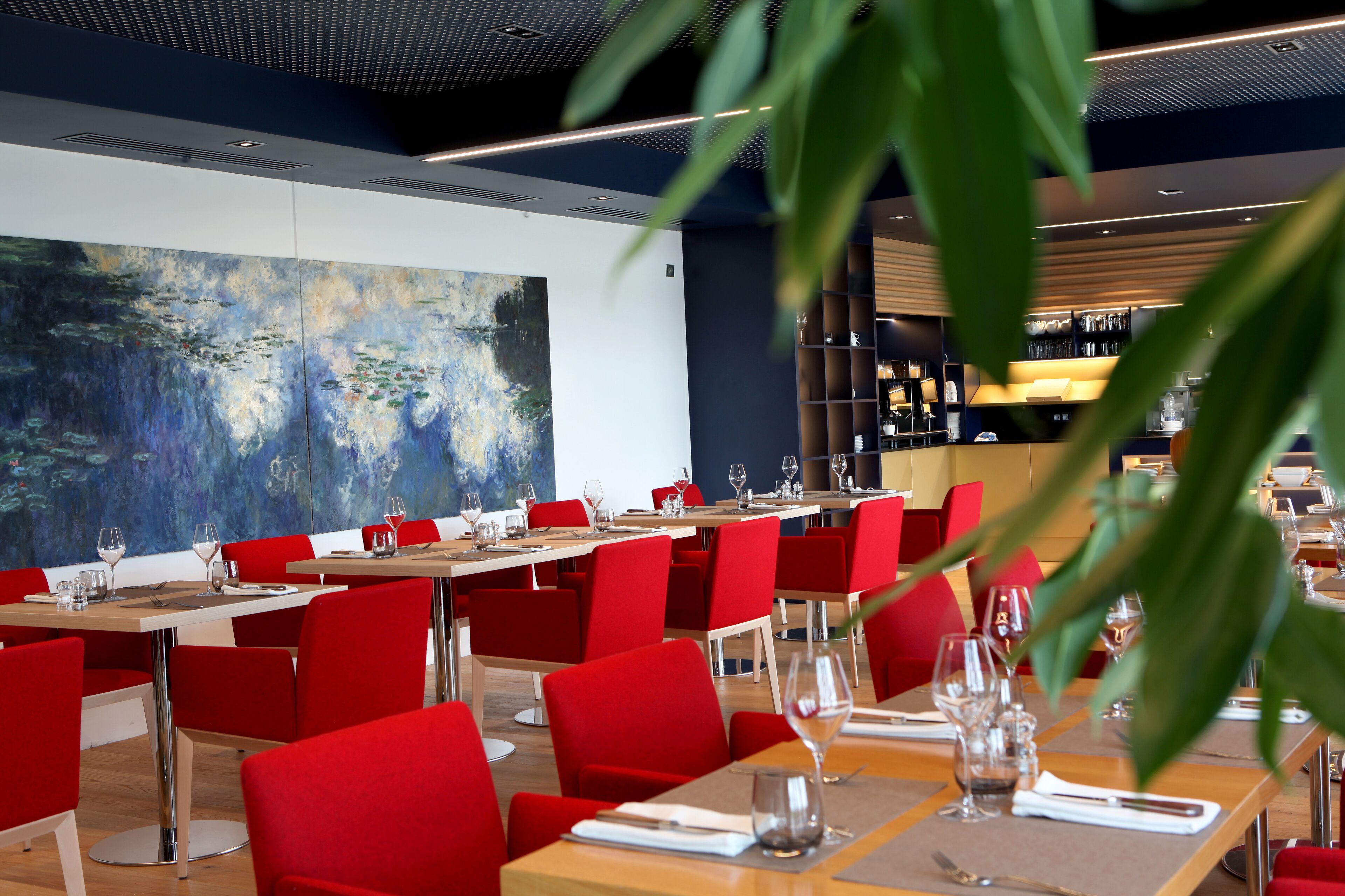 A restaurant with red chairs and tables, a large painting on the wall, and a modern ceiling design.