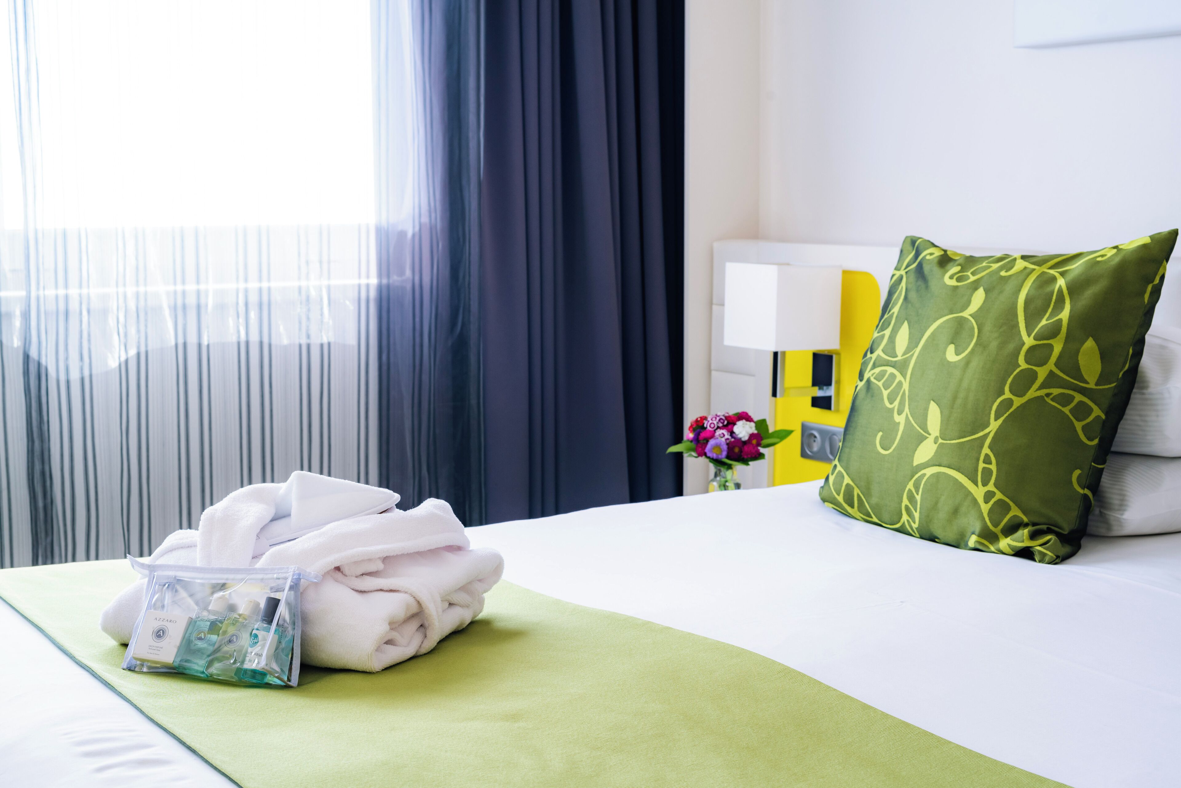 A neatly made bed with a green bedspread, a pillow with a leaf pattern, and a small bag of toiletries on the side.