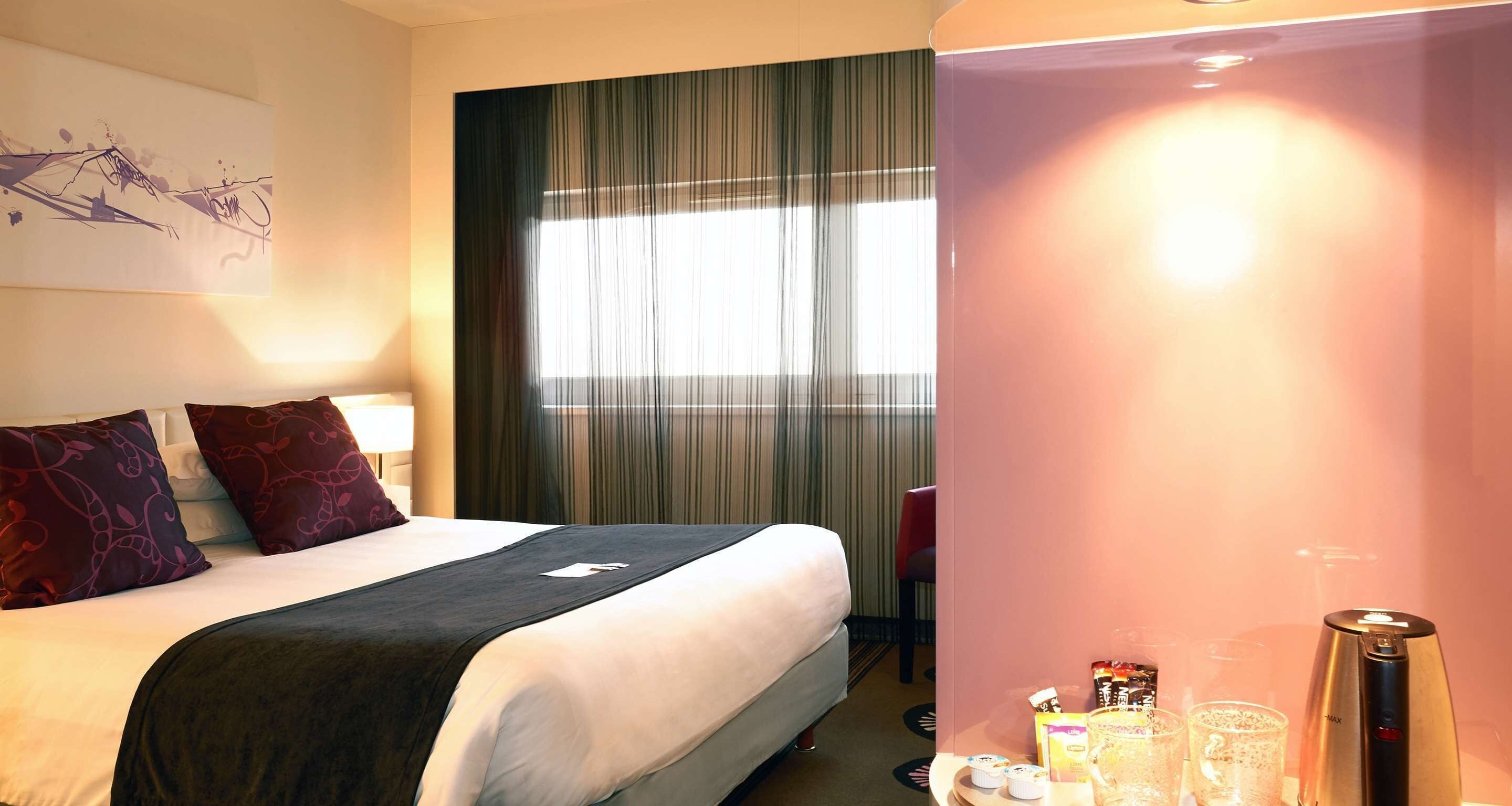 A hotel room with a bed, a bedside table with a coffee maker, a window with curtains, and a wall with a painting.