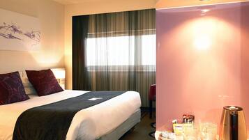 A hotel room with a bed, a bedside table with a coffee maker, a window with curtains, and a wall with a painting.