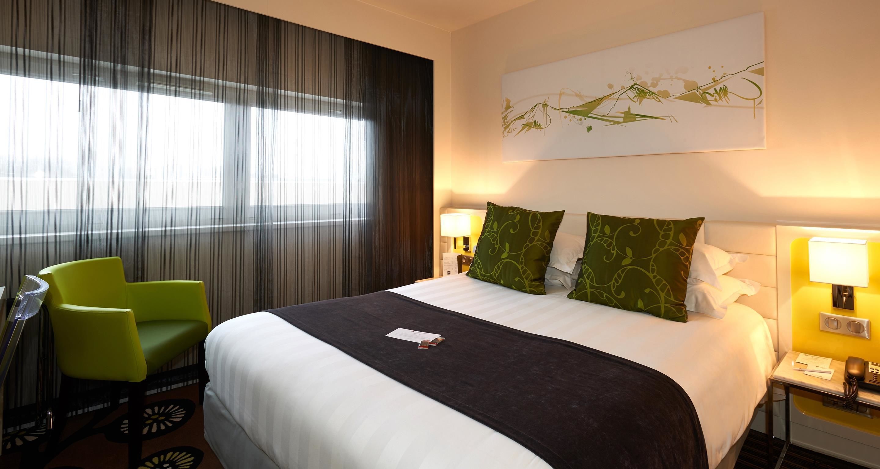 A hotel room with a bed, green chairs, a desk, and a lamp.