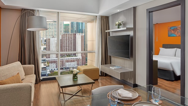 A modern hotel room with a large window, a sofa, a dining table, and a bed. - F1 Hotel Manila (Taguig)