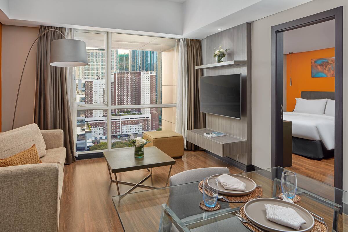 A modern hotel room with a large window, a sofa, a dining table, and a bed.