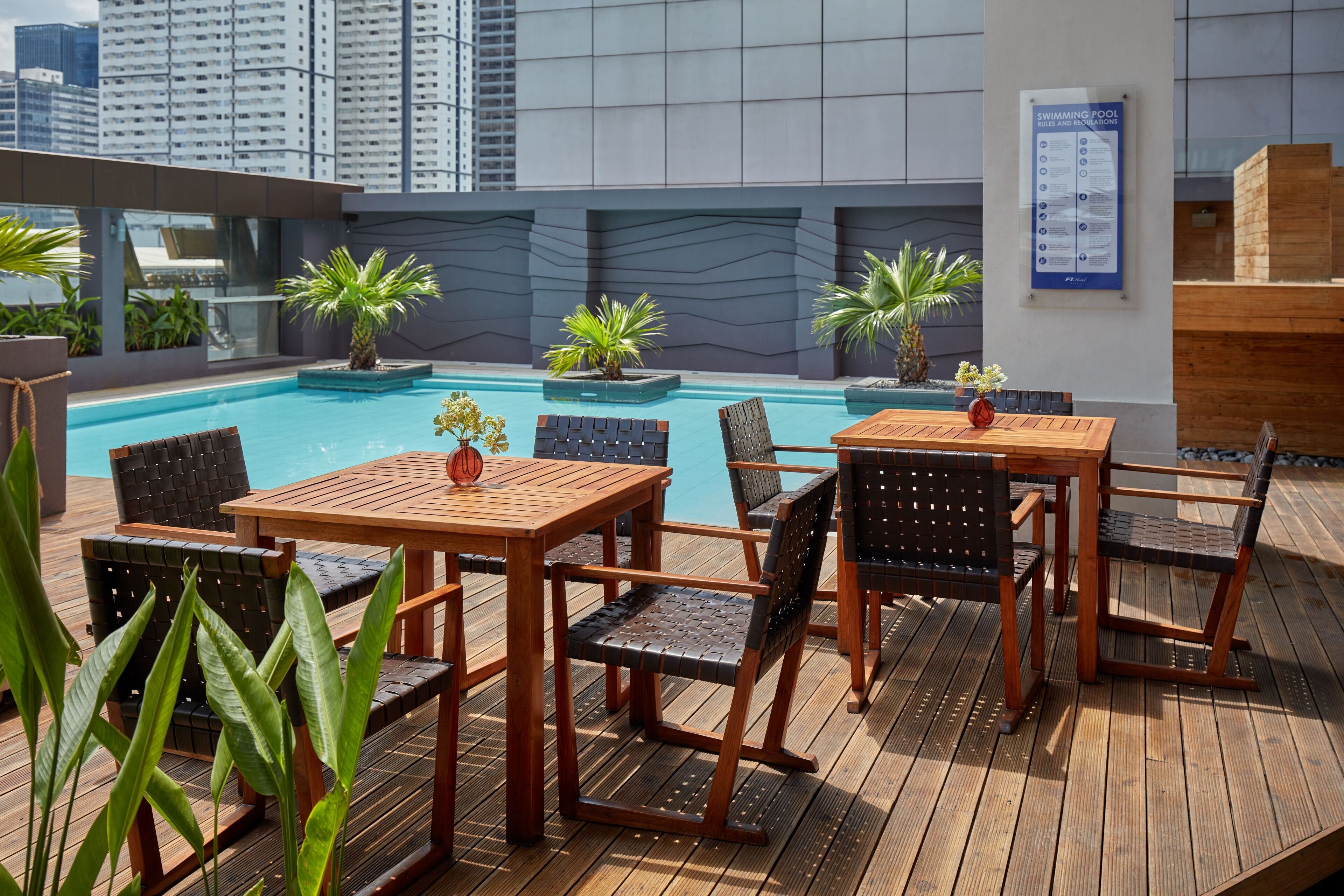 A rooftop pool area with wooden tables and chairs, surrounded by potted plants.