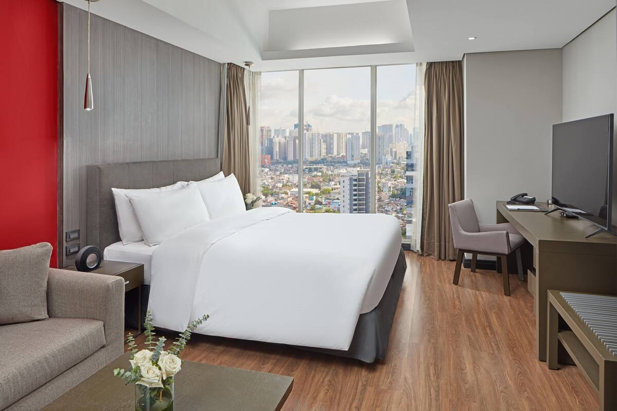 A modern hotel room with a large bed, a desk, and a view of the cityscape.