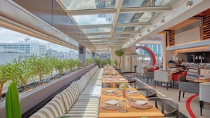 A rooftop restaurant with a long bench seating area, tables with place settings, and a view of the cityscape through a glass ceiling. - F1 Hotel Manila (Taguig)