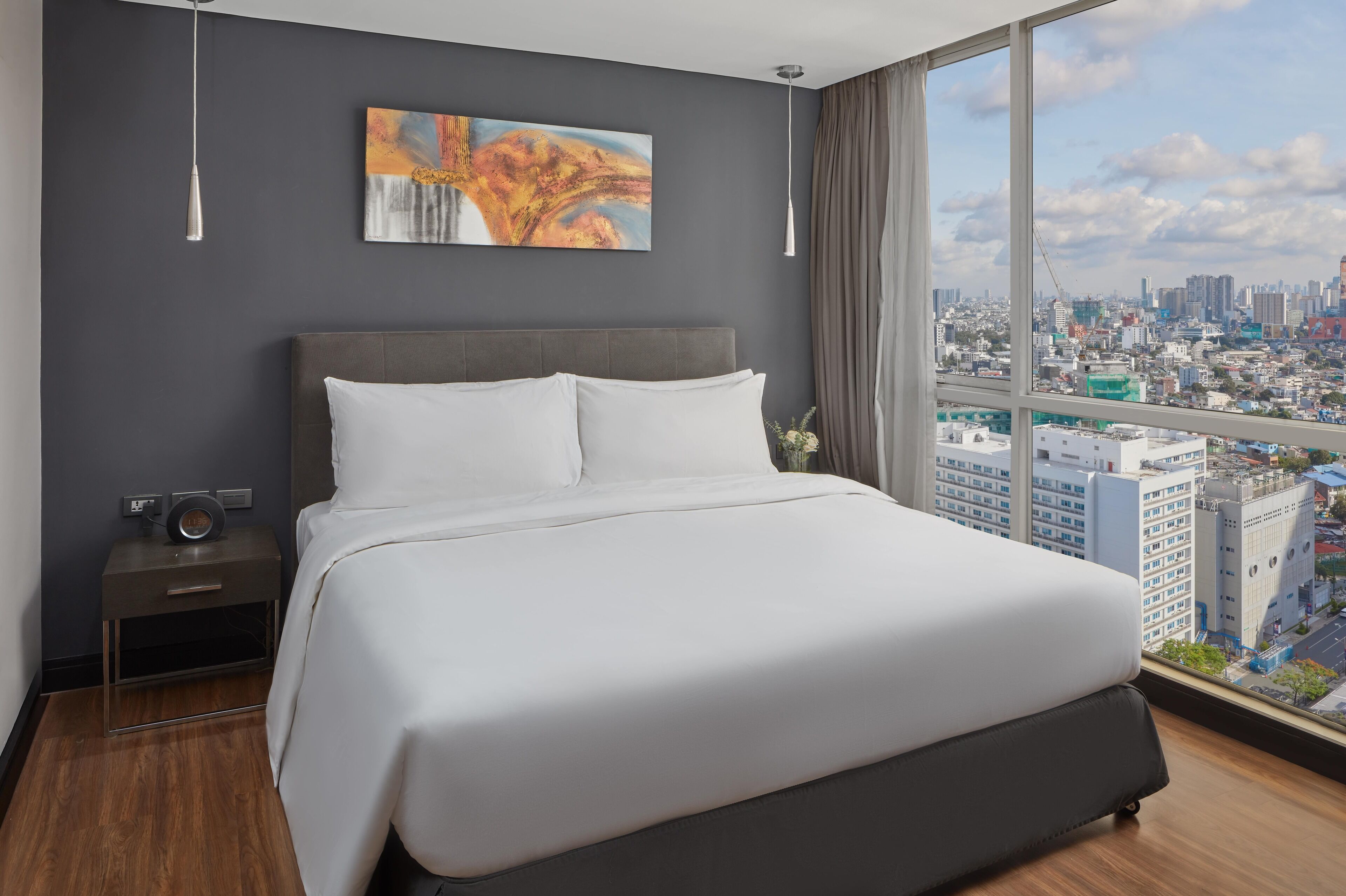 A modern hotel room with a large bed, a nightstand, a painting on the wall, and a city view through the window.