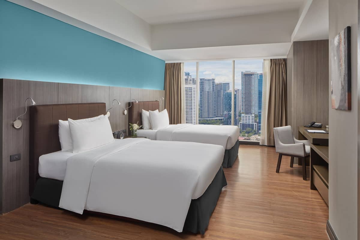 A hotel room with two beds, a desk, and a view of the cityscape.