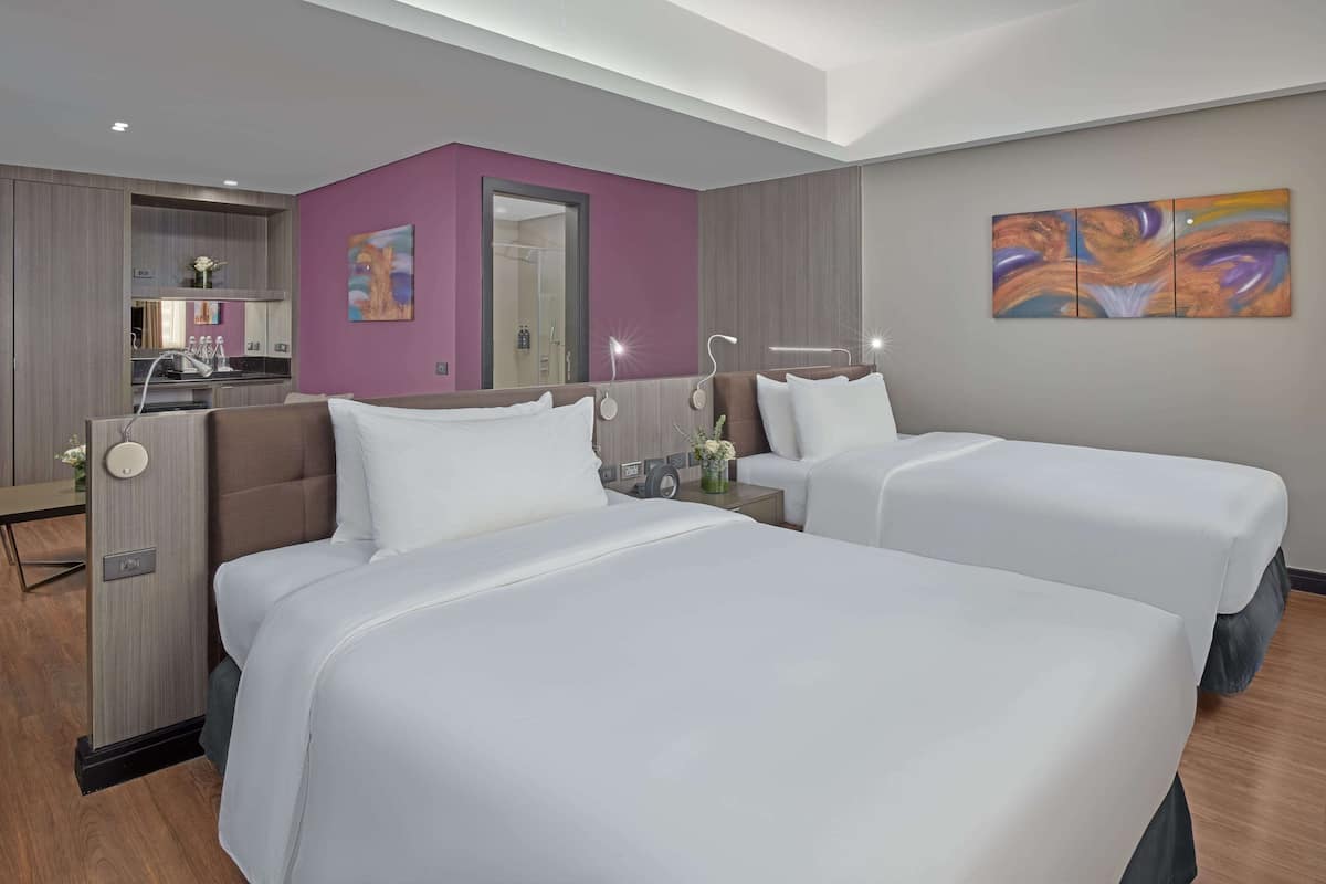 A hotel room with two beds, a kitchenette, a bathroom, and a large painting on the wall.