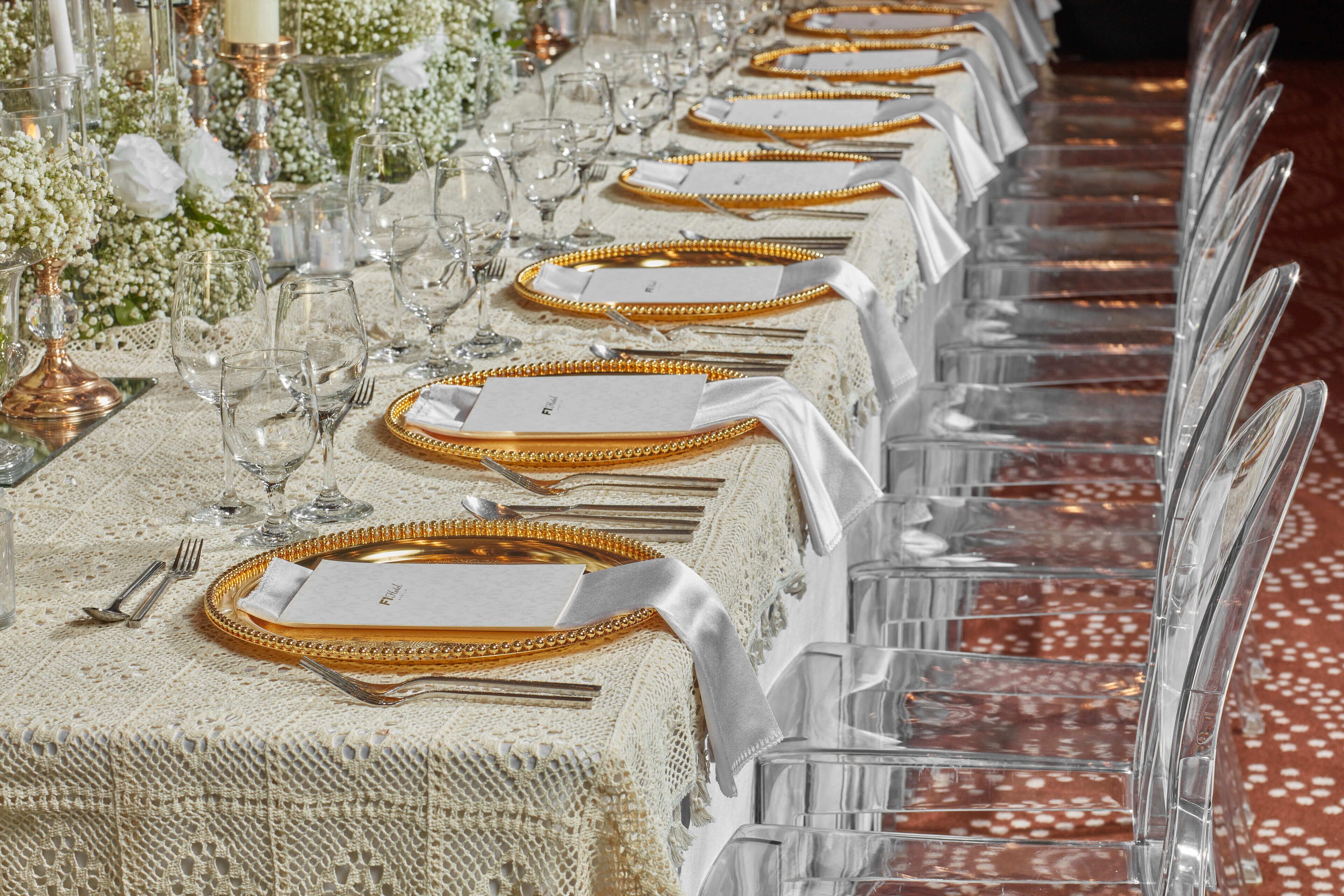 Una larga mesa de comedor dispuesta con platos de borde dorado, copas de cristal transparente y servilletas blancas.