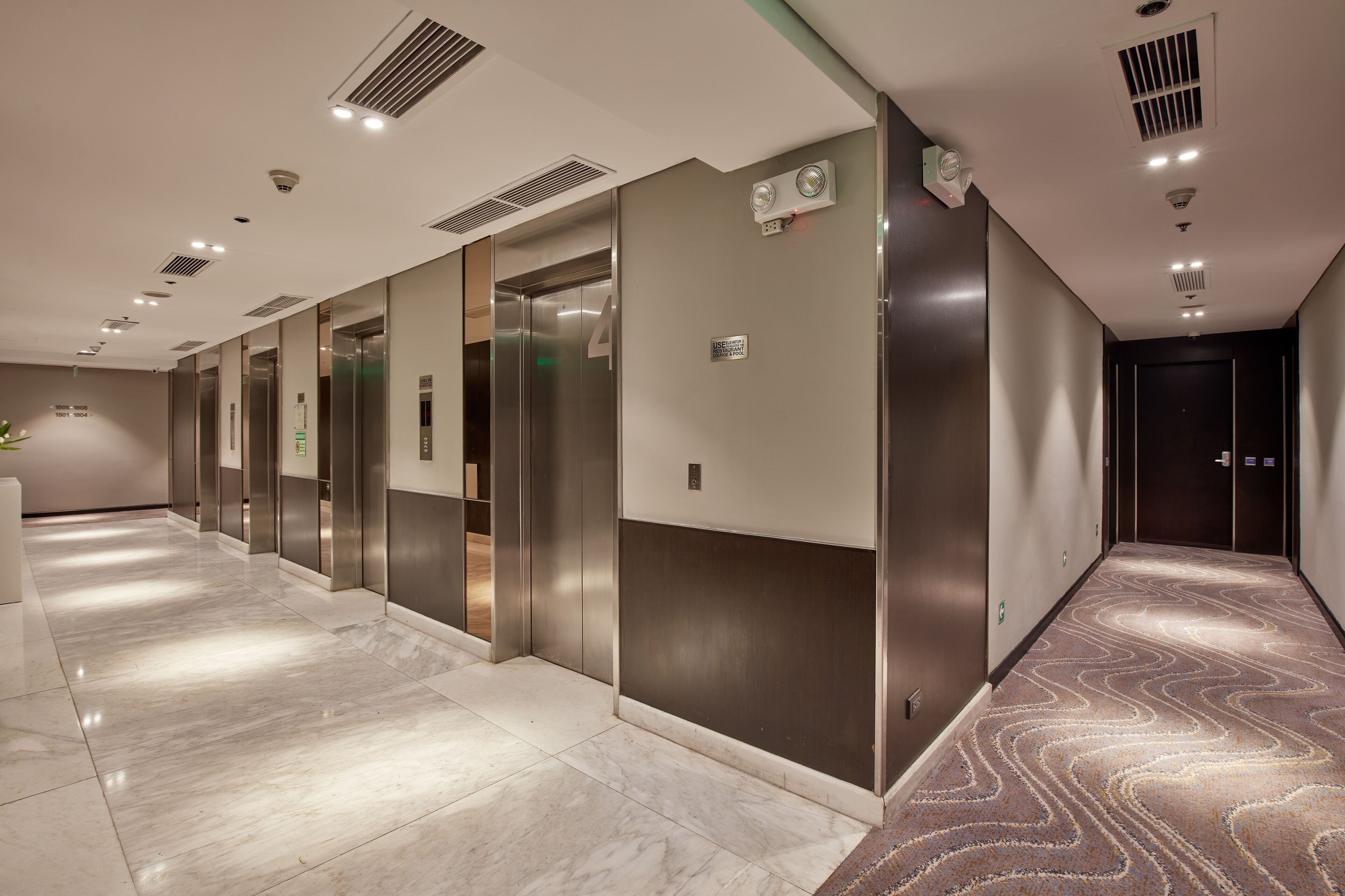 A modern hotel corridor with multiple elevators, marble flooring, and a patterned carpet.