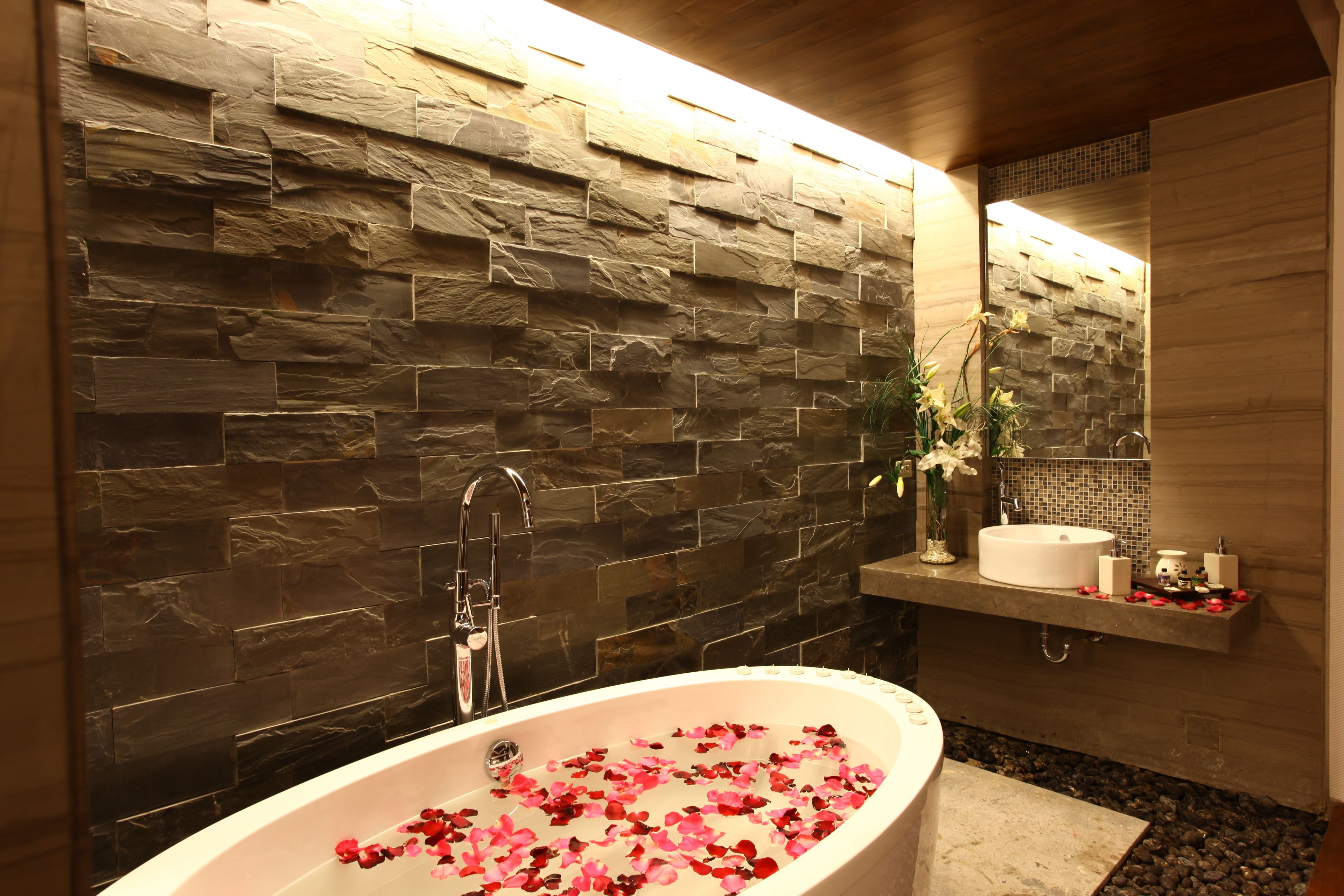 A modern bathroom with a stone wall, a freestanding bathtub filled with rose petals, a sink, and a mirror.