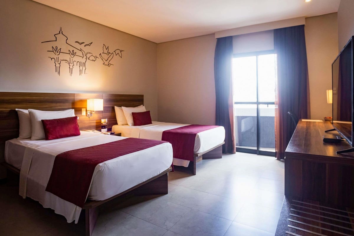A hotel room with two beds, a TV, a desk, and a large window with curtains.