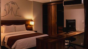 Habitación de hotel con cama, escritorio, silla, lámpara, televisor y armario.