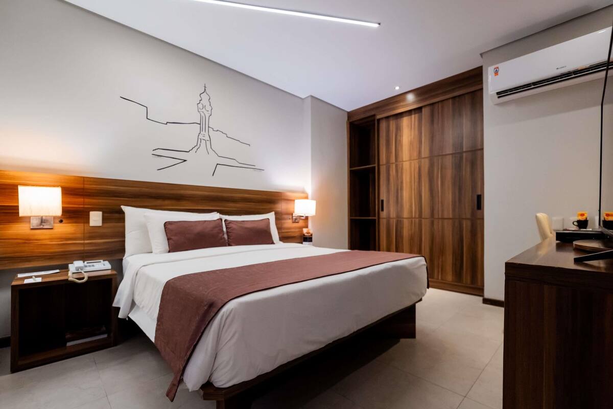 A modern hotel room with a large bed, wooden headboard, bedside lamps, a desk, and a wardrobe.