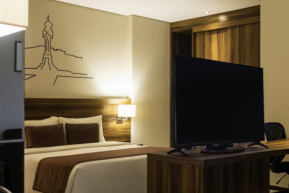 Quarto de hotel com cama, escrivaninha com televisão, mesa de cabeceira com abajur e um objeto decorativo na parede.