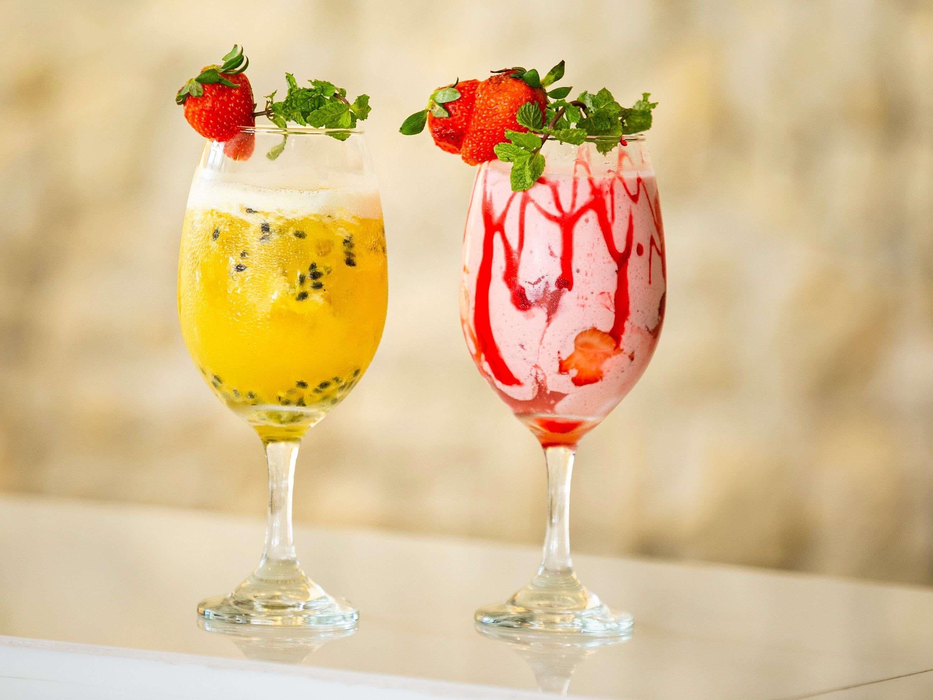 Two cocktails in stemmed glasses, one with a yellow base and the other with a red base, both garnished with a strawberry and mint leaves.