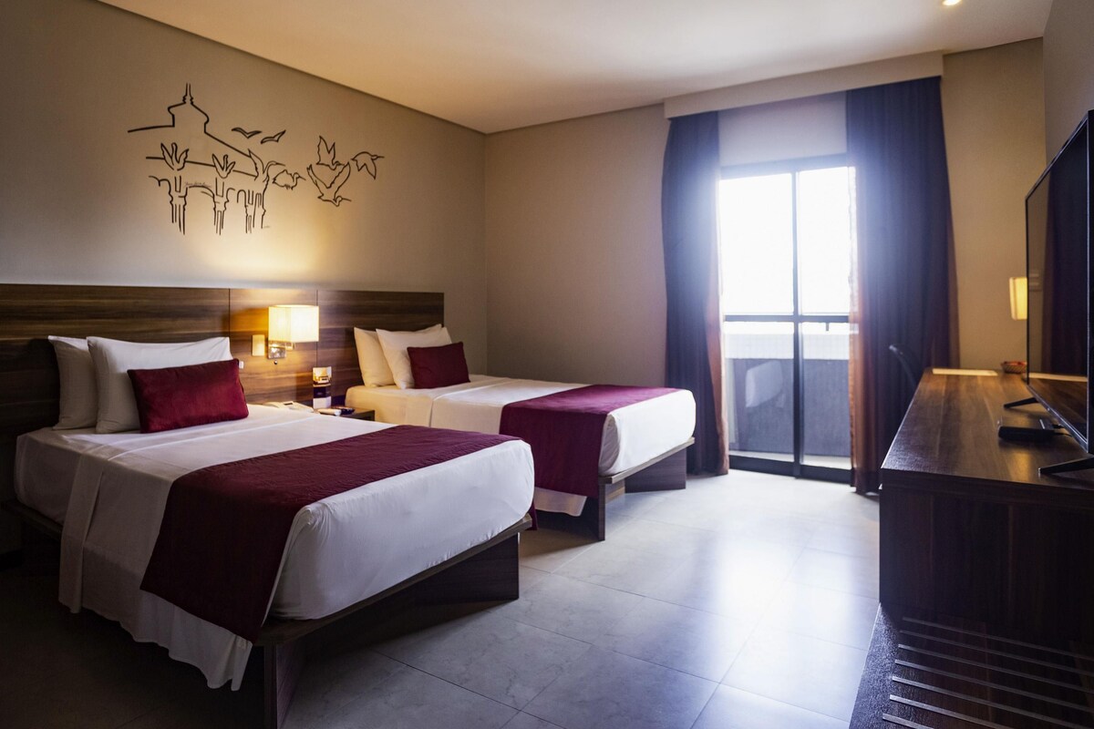 A hotel room with two beds, a TV, a desk, a window with curtains, and a wall art piece.