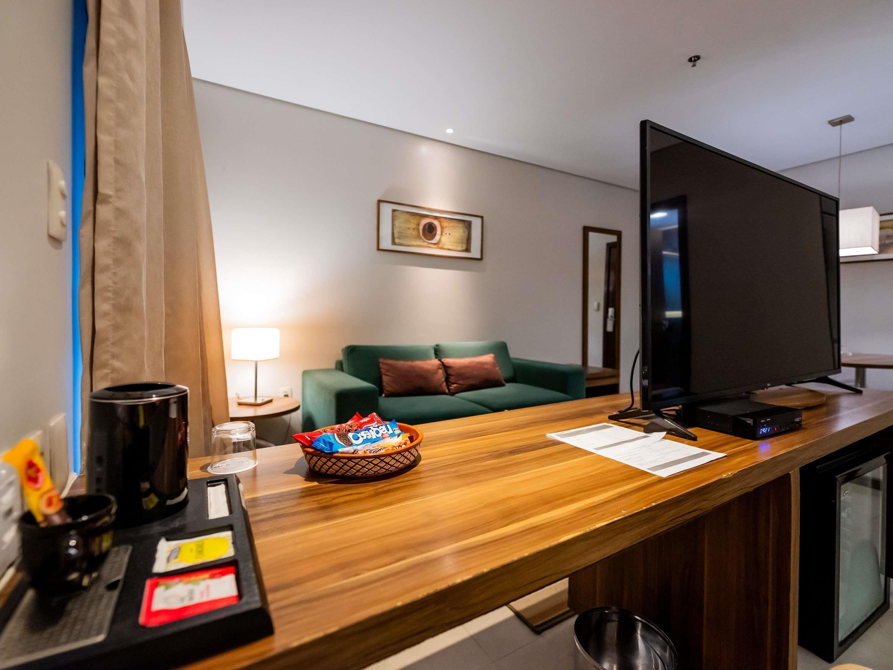 Habitación de hotel con escritorio de madera, televisor de pantalla plana, sofá verde, lámpara y un cuadro enmarcado en la pared.