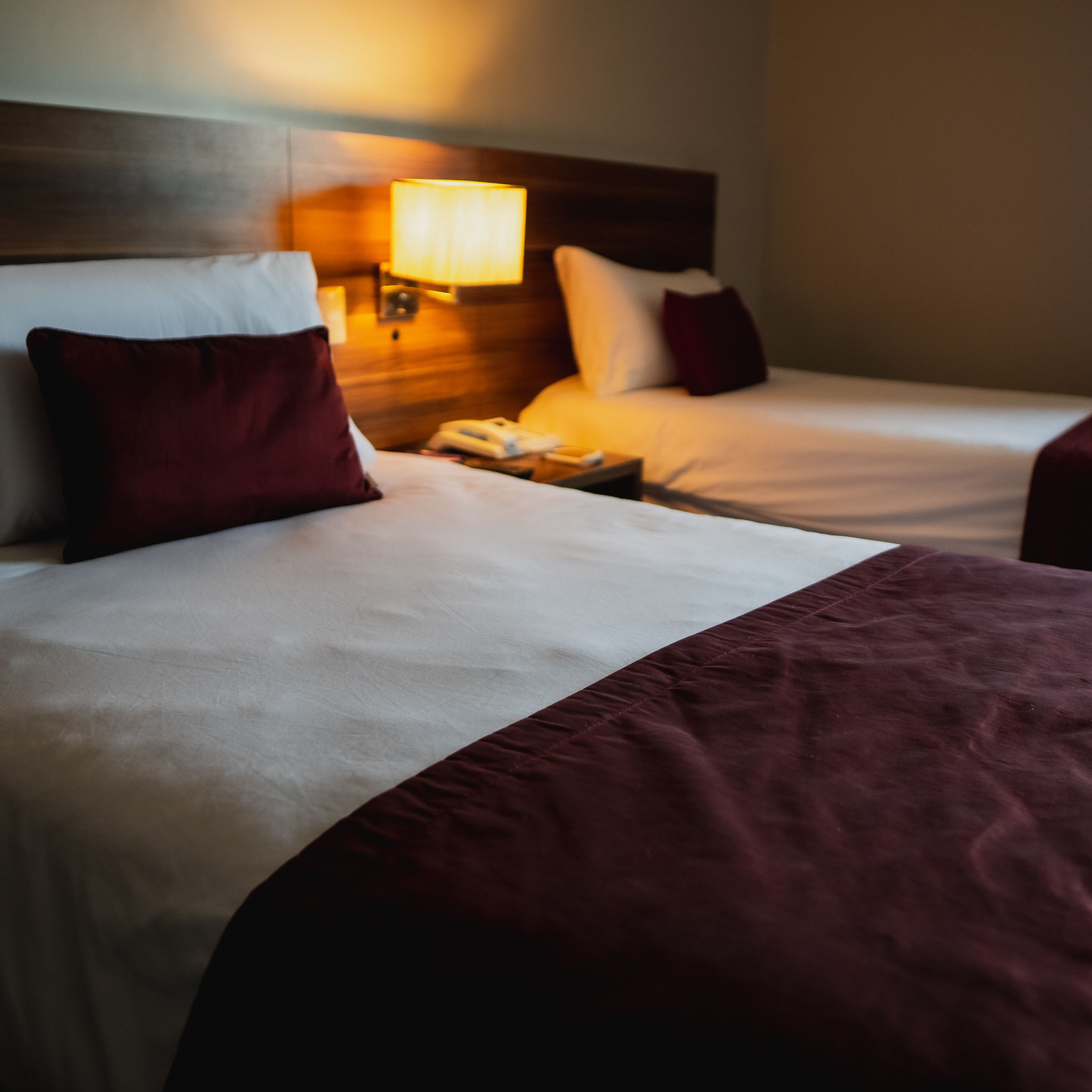 Habitación de hotel con dos camas, cada una con una colcha blanca y burdeos, un cabecero de madera y una lámpara de noche.