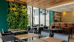 A modern restaurant interior with glass tables, black chairs, a green wall with plants, and a stone wall bar area. - Mercure Belém Boulevard (Belém)
