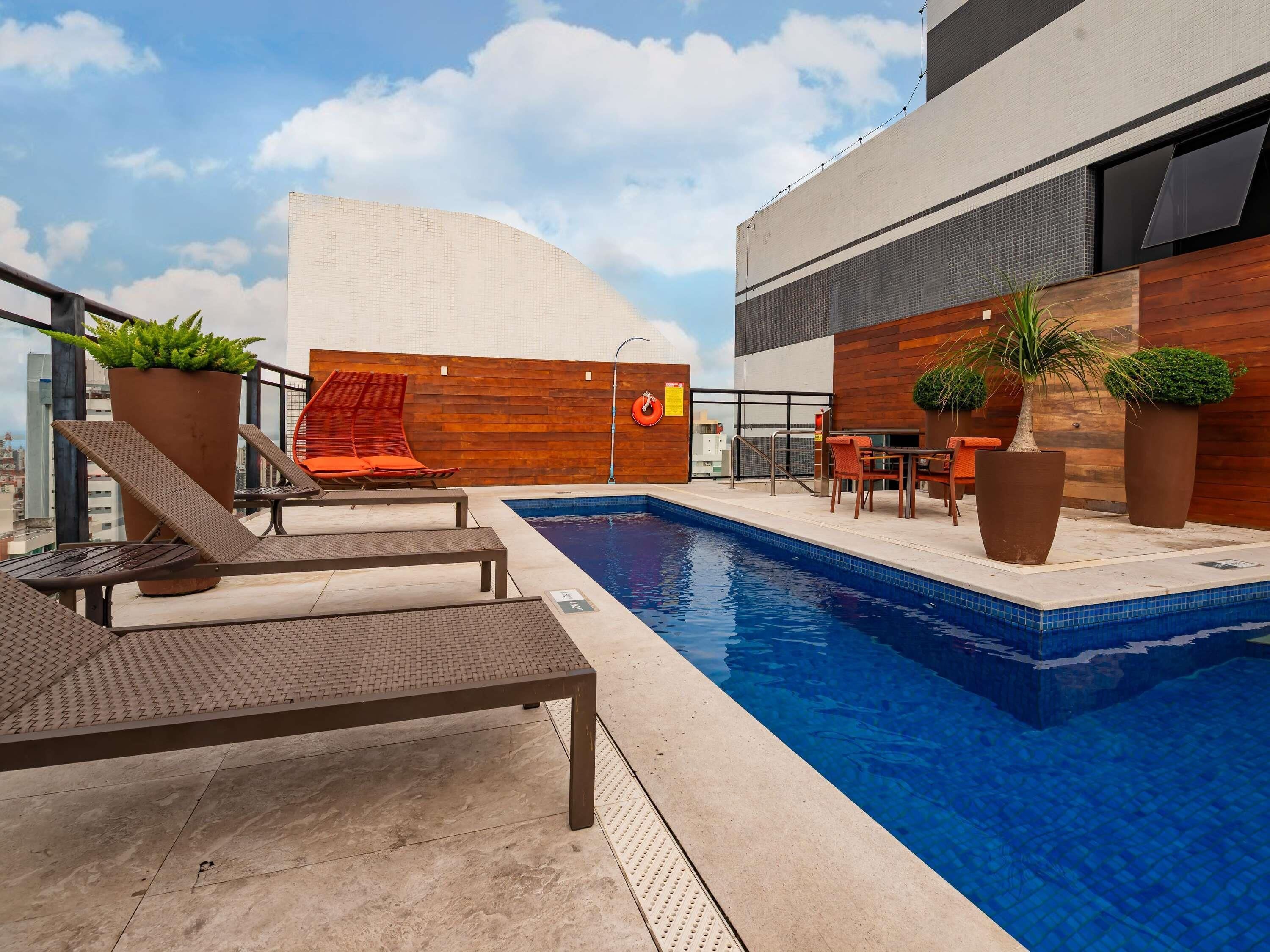 Uma área de piscina no terraço com espreguiçadeiras, uma mesa e plantas em vasos.