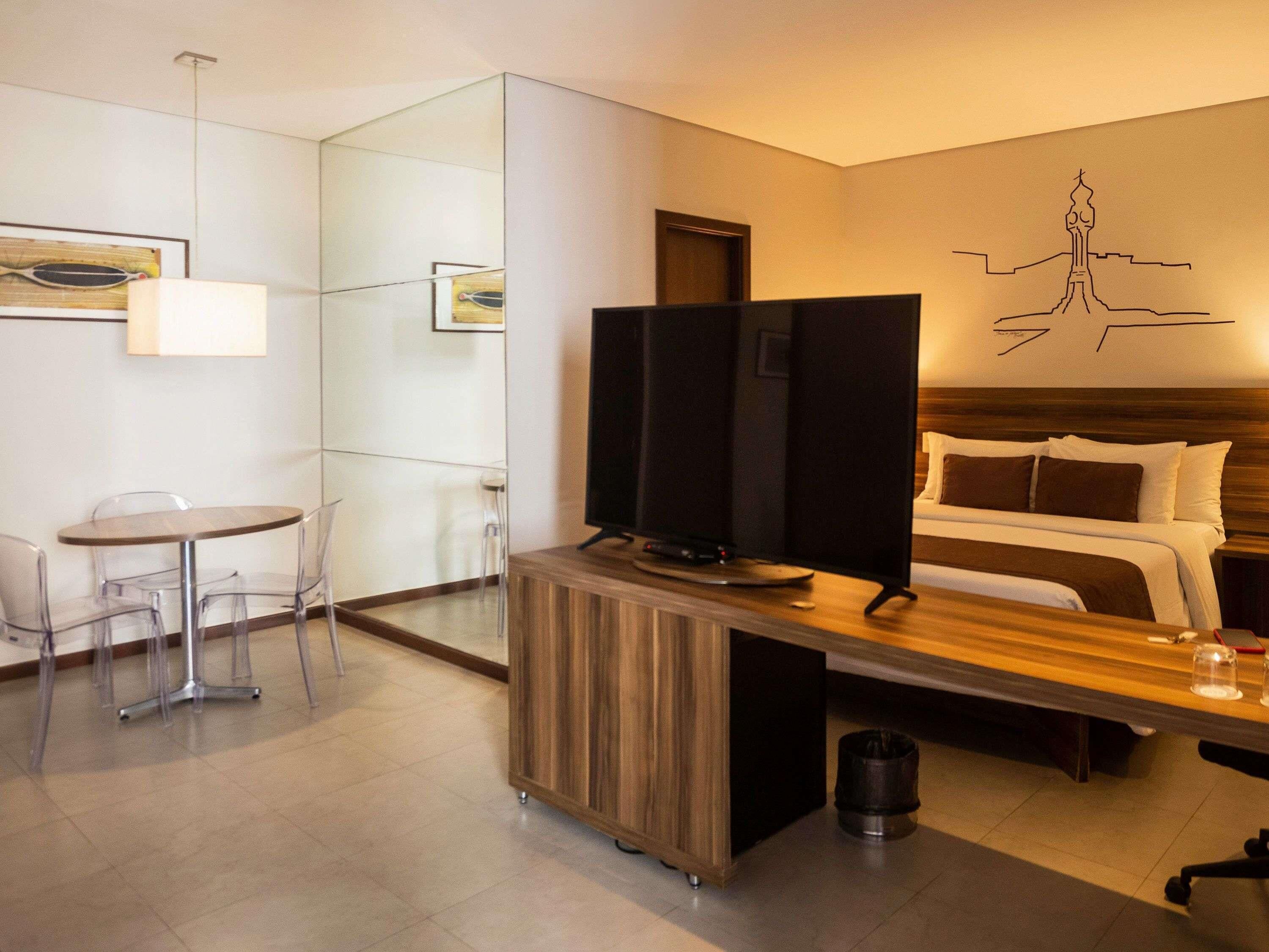 Ein Hotelzimmer mit einem Bett, einem Schreibtisch mit Fernseher, einem kleinen Tisch mit Stühlen und einer Wand mit einer schlichten Zeichnung.
