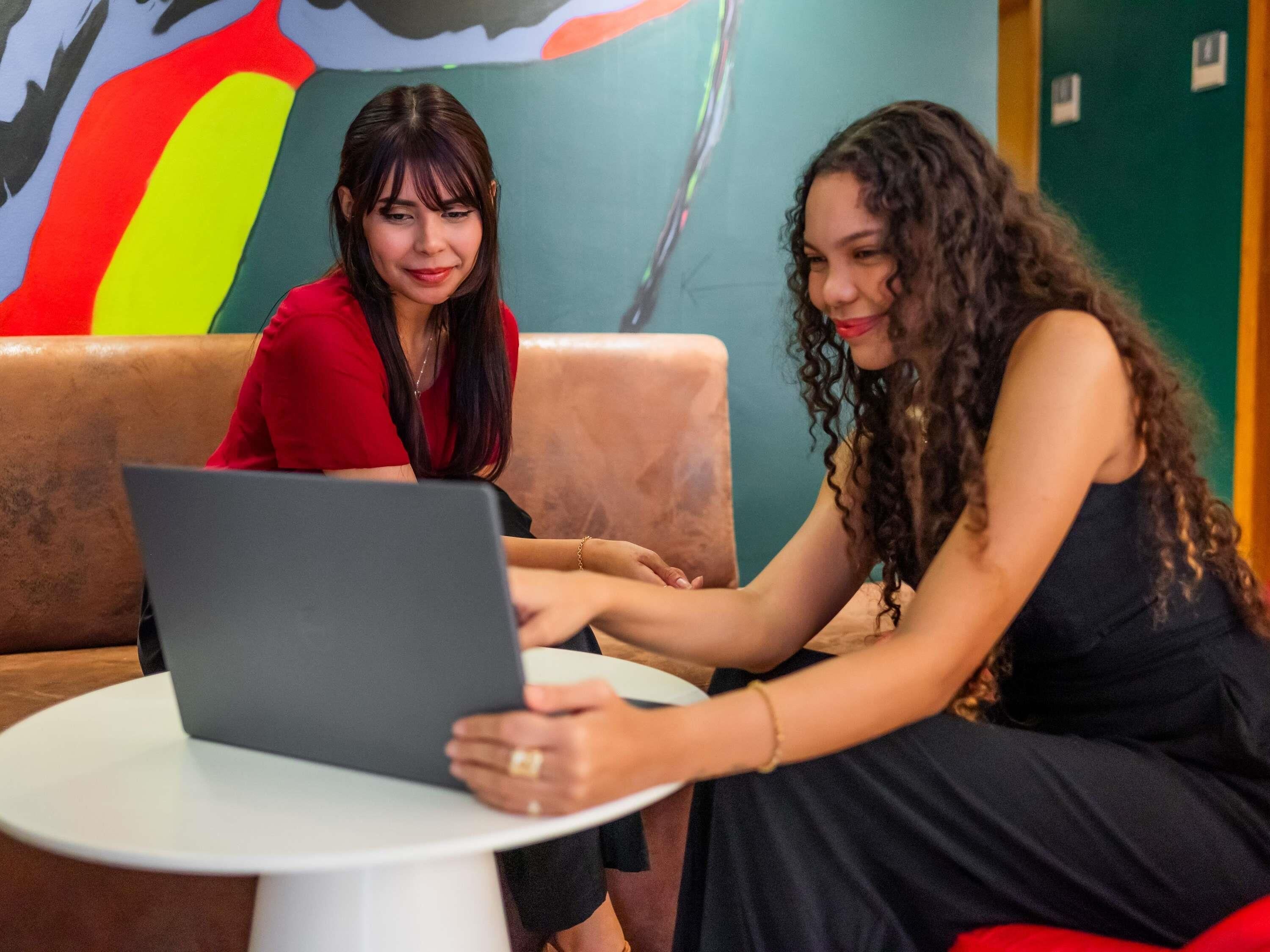 Two individuals seated on a couch with a laptop on a small table, in a room with a colorful mural on the wall.