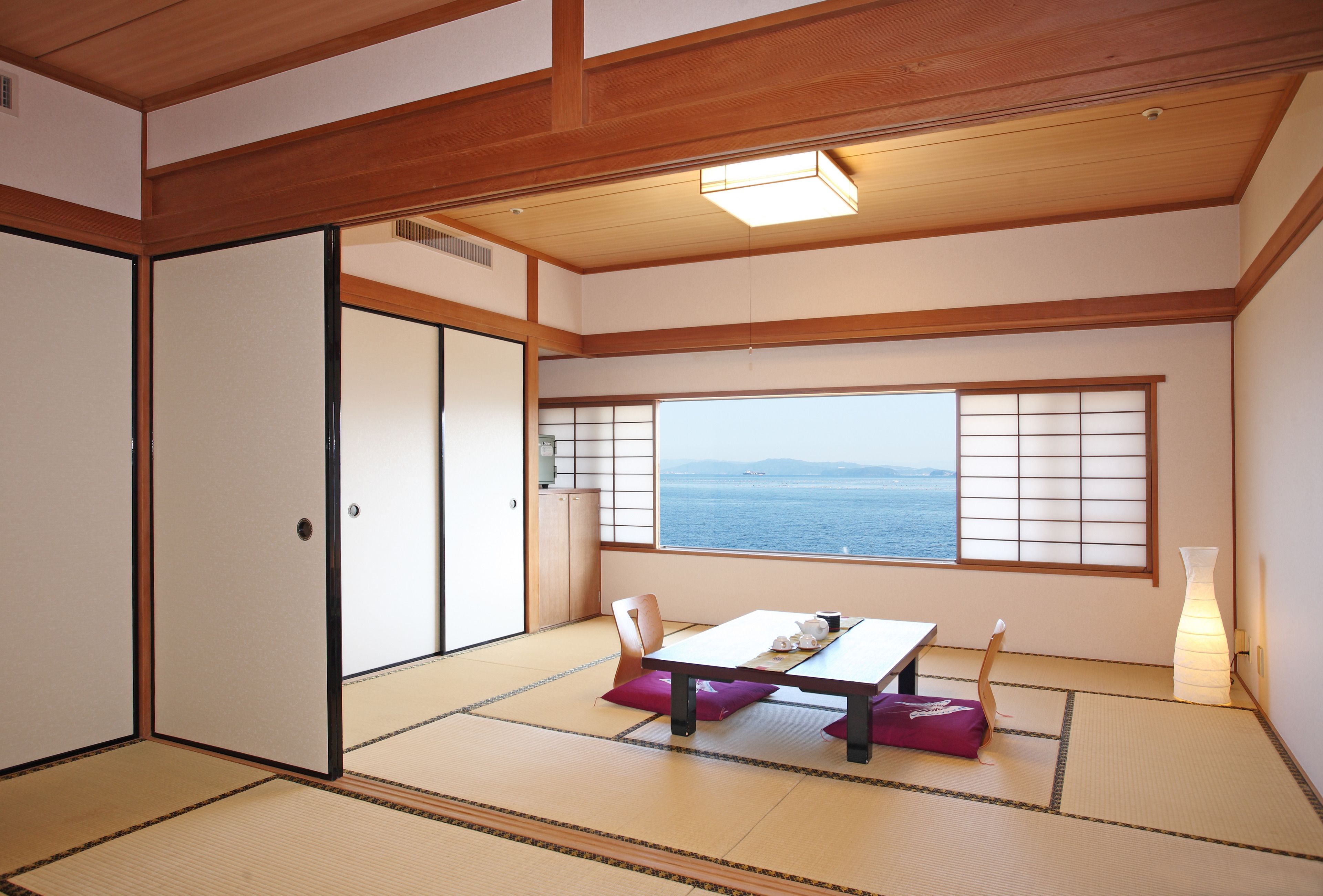 A traditional Japanese room with tatami flooring, a low table with cushions, a sliding door, and a view of the sea through a shoji screen.