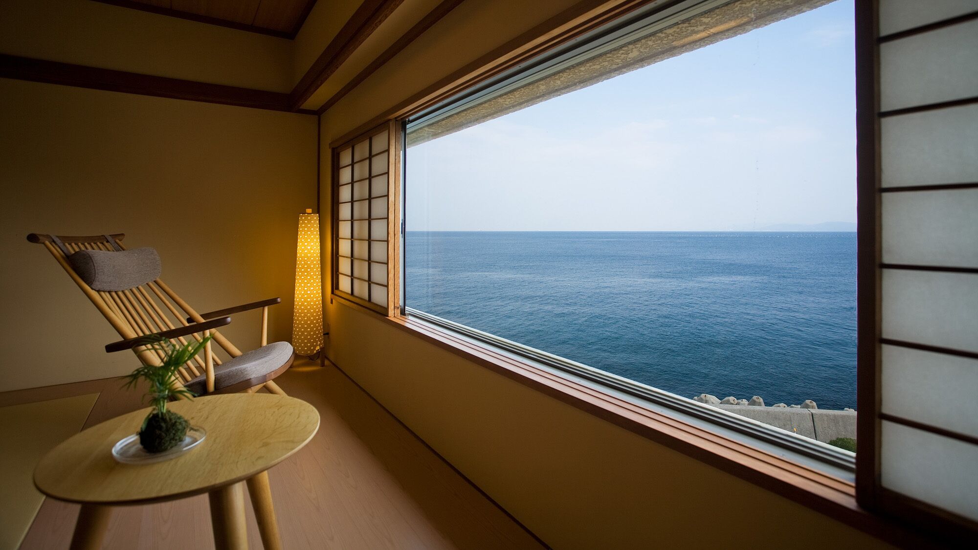 A room with a view of the sea, a wooden chair, a small table with a plant, and a traditional Japanese lantern.